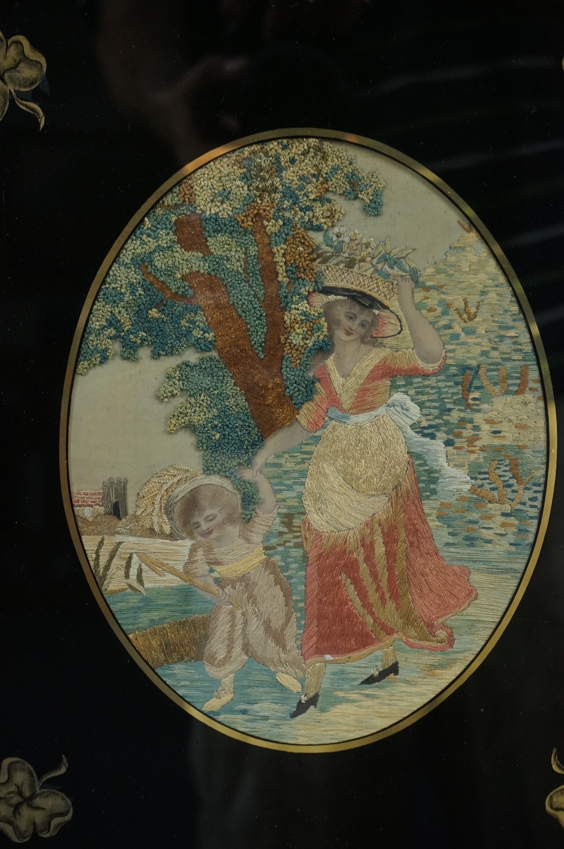 Two early 19th century oval silk worked embroideries. Both figural pastoral scenes, mostly worked in stem stitch, the leaves in a stump work style knotting. Both embroideries set within decorative Verre Eglomise glass an